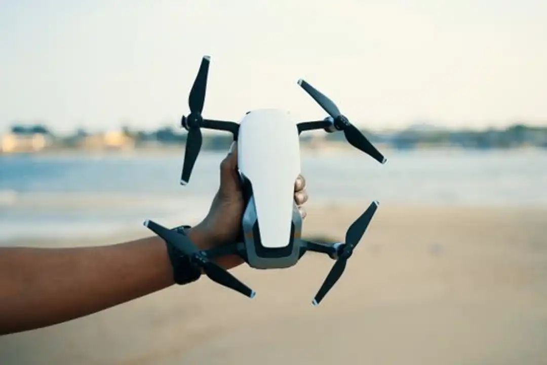 Tangan memegang drone modern di pantai saat siang hari.