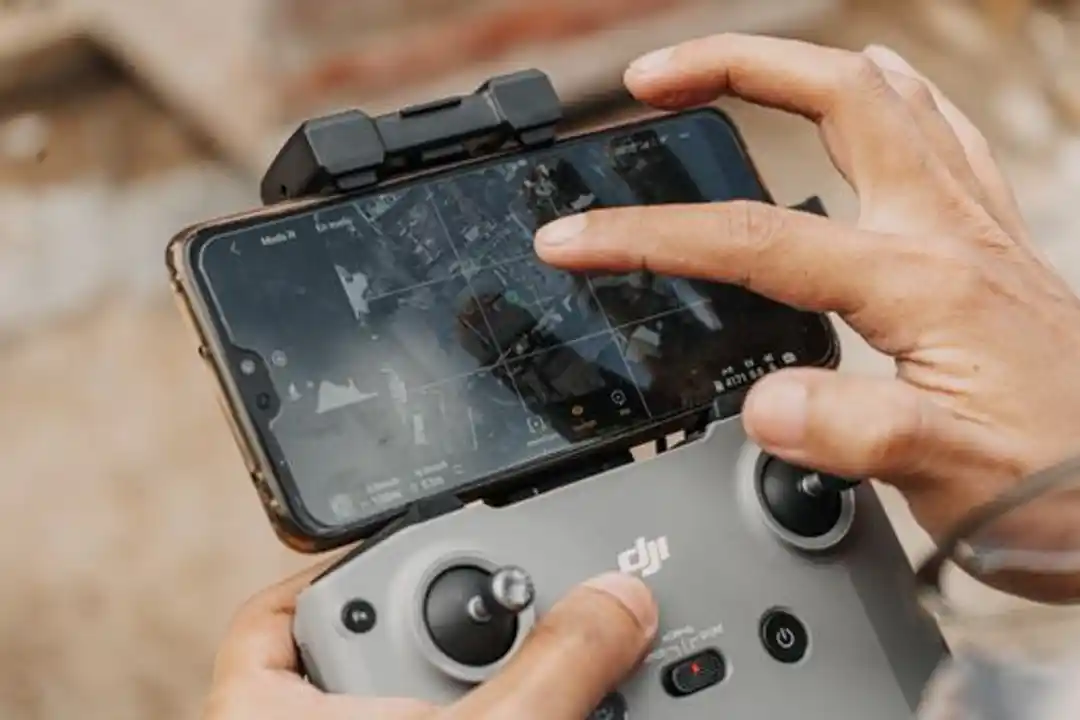 Tangan memegang kontroler drone DJI dengan smartphone, menampilkan teknologi navigasi.