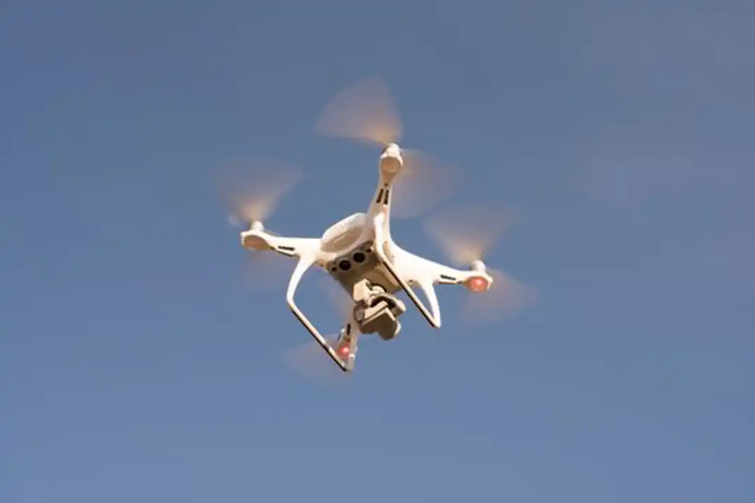 Drone putih terbang di langit biru cerah, merekam rekaman udara dengan presisi.